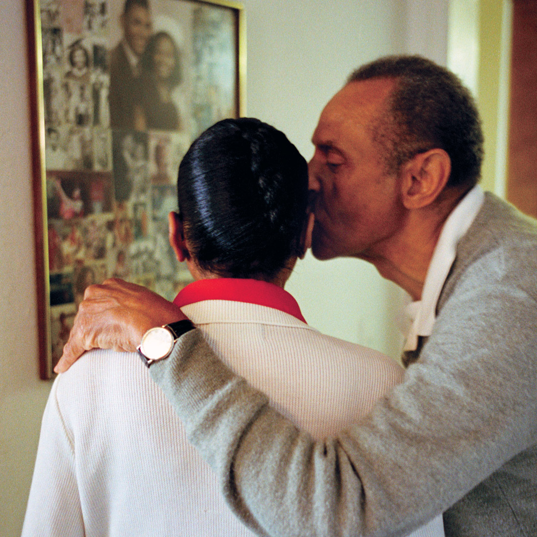 man kissing wife on cheek with family photos hanging on wall in background